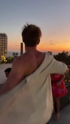 The video shows a man from behind wearing a white shirt and dark shorts. He is standing on a balcony with a railing. In the background, there is a sunset with orange and yellow hues. To the right, there is a woman in a pink and red dress. She is also on the balcony and appears to be talking to the man. There are trees and some buildings visible in the distance. The man is turning his head and looking towards the woman.