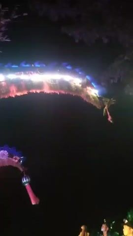 This video captures a lively night scene in Dukuh Klopo, Indonesia, likely during a festive event. The primary subjects are a group of people gathered around a large, elaborate sound system. The sound system is adorned with a vibrant archway, decorated with what appear to be traditional festive ornaments, possibly related to a harvest or religious celebration, and numerous colorful lights, including blue, white, green, and red LEDs.

The people are dressed in casual attire, with some wearing matching t-shirts featuring a distinctive tiger emblem and the word "Aljabar". Their body language suggests engagement with the event, with some looking towards the sound system, others interacting with each other, and a few appearing to be dancing or reacting to music. The presence of a person on the ground, possibly sitting or lying down, adds to the dynamic and perhaps informal nature of the gathering.

The setting is outdoors at night, indicated by the darkness and the prominent use of artificial lighting from the sound system and string lights. The backdrop includes trees and foliage, hinting at a rural or semi-rural location. A flag, possibly the Indonesian flag given its red and white colors, is visible on a pole behind the archway.

The activity appears to be a musical performance or a celebration where music and lights are central. The energy conveyed through the flashing lights and the people's reactions suggests a festive and celebratory atmosphere. There is some visible text on the side of the speakers, but it is largely obscured or illegible due to the low resolution and lighting conditions. The overall impression is one of a community event filled with music, light, and communal enjoyment.
