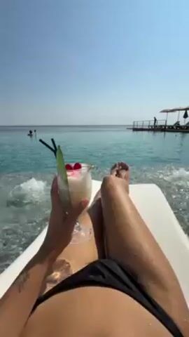 A person in a black bikini lies on a white lounger, holding a tropical cocktail with a straw and cherry garnish. The cocktail is white and appears to be a frozen drink. The person has tanned skin and is facing a turquoise body of water that appears to be the ocean, under a clear blue sky.  There are people swimming in the distance and a dock with umbrellas and people visible. Small waves splash onto the lounger. The scene is bright and sunny, giving the impression of a relaxing tropical vacation.