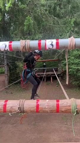 The video shows a person navigating an obstacle course in a forest. The individual is wearing a helmet and a harness, attached to safety lines, and is carefully walking across a suspended log. They are surrounded by trees, suggesting an outdoor adventure park or similar recreational facility. Another person can be seen in the background on a wooden platform. The scene appears to be taking place during daylight hours, with lush green foliage indicating it's likely spring or summer. The overall mood is one of challenge and focus as the person concentrates on their balance. The log they are crossing has red markings on it, and a higher pipe structure also has visible numbers "2" and "3" marked on it, possibly indicating different stages or parts of the course. The ground below is dirt, consistent with a trail or natural terrain. The focus is on the physical activity of traversing the obstacle, highlighting the skill and concentration required.