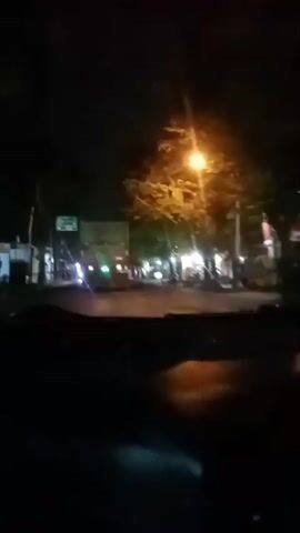The video, filmed at night from the interior of a vehicle, captures a view of a street. In the foreground, the dashboard and windshield are visible, with streaks of light reflecting off the glass. The primary focus is a large, light green truck with a white cargo box, seen from the rear. Its red taillights are illuminated, and there are green lights visible on the rear of the cargo box. The truck is in the left lane of a road. To the right of the truck, the road continues, with several trees lining the side. Streetlights cast bright, diffused illumination, creating lens flare effects. Several figures, appearing to be people, are standing near the right side of the road, some by what appear to be stalls or shops with lights. There are also motorcycles parked along the right side. The overall scene suggests a moderately busy street at night, possibly a marketplace or commercial area.