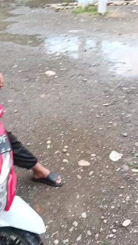 The video shows a group of young children playing in a muddy puddle on a dirt road. The children are wearing what appear to be school uniforms. One child in a white shirt and pants stands in the puddle, while another child in a similar uniform kneels in the mud, playing with his hands. Two other children are also in the puddle, one in a white shirt and pants, and another in a darker shirt and pants. In the background, there are some buildings and trees, and other children can be seen walking further down the road. The overall environment appears to be a rural or semi-rural area.