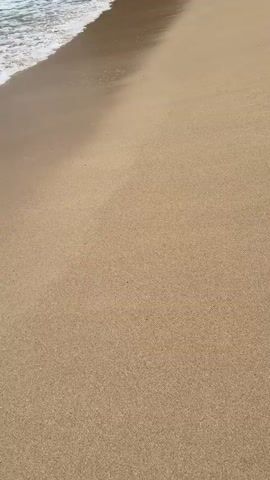 The video shows a beach from a high angle. A thin layer of ocean water with white foam rolls onto the sand. The sand is light brown and mostly clear, except for a few scattered small objects. The camera is stationary, focused on the meeting point of the water and sand. The footage is clean and well-lit.