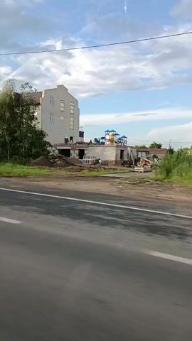 The video shows a street view from a moving vehicle. On the left side of the street, there is a multi-story building with dormer windows. A tall lamppost stands in front of the building. To the left of the building, there is a road sign with blue directional arrows. The area around the building is covered in green grass and trees. Further down the street, there appears to be a construction site with building materials and a playground structure. The sky is partly cloudy. The road in the foreground has white lane markings. The video captures a passing view of this scene.