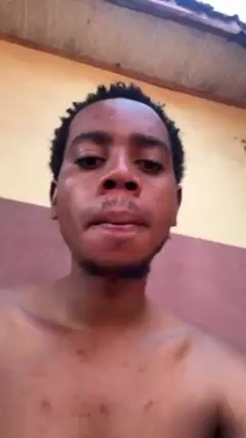 The video shows a shirtless young man looking directly at the camera. He has short, curly hair and a light beard. His skin appears slightly reddish, possibly sunburned, and he has a few noticeable blemishes or marks on his chest. He stands in front of a wall that is painted in two colors: a creamy yellow at the top and a reddish-brown at the bottom. A wooden ceiling or overhang is partially visible at the top of the frame. The man's facial expression is neutral.