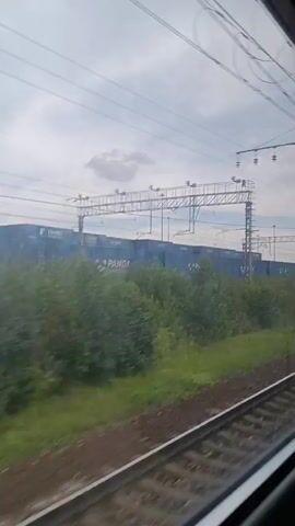 The video shows a view from a moving train window. Outside the window, there are train tracks, grassy areas, and trees. In the background, there are blue shipping containers stacked together. There are also power lines and poles overhead, indicating an electrified railway line. The sky is cloudy.