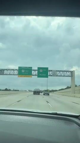 The video shows a view from inside a vehicle driving on a highway. The sky is overcast with a lot of clouds. There are two green highway signs visible. The sign on the left says "Express Lane 290 EAST 1 1/4 Miles Left Entrance". The sign on the right says "290 EAST Houston". There is also a speed limit sign that says "SPEED LIMIT 65". Several vehicles are on the highway, including cars and a truck. The highway has multiple lanes. The scene appears to be in Texas given the mention of Houston.