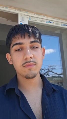 A young man stands outside, taking a selfie-style video. He has dark hair, brown eyes, and a small mustache and goatee. He wears a dark blue button-down shirt with short sleeves. The camera pans down to show him wearing black athletic shoes while sitting on a rough concrete surface with brick debris and an orange extension cord nearby. The background shows a glass door and a glimpse of the outdoors with a blue sky.
