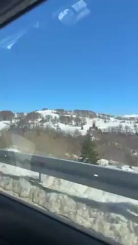 The video is taken from inside a moving vehicle. The camera is pointed out the window at a snowy landscape. The sky is clear and blue. There are hills in the distance covered in snow. There are trees and a few buildings visible on the hills. In the foreground, there is a metal guardrail with snow accumulated next to it. The overall impression is of a sunny winter day in a mountainous area.