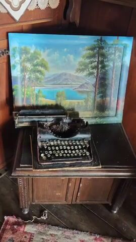 The video opens with a panning shot of a vintage typewriter on a wooden desk, with a landscape painting hanging above it. The camera then moves to a close-up of a framed portrait of a man with a prominent mustache. The scene shifts to a wall with a framed document and a shelf of old books. The lighting is dim, suggesting an indoor setting, possibly a museum or historical exhibit. The overall impression is one of a preserved historical space.