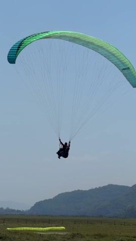 A paraglider is landing in a grassy field on a sunny day. The paraglider is green and blue and has the word "ADCO" on it. A person is harnessed to the paraglider and holding onto the control lines. There are two people standing in the field, waiting for the paraglider to land. A small body of water is at the edge of the field. In the background is a mountain range.