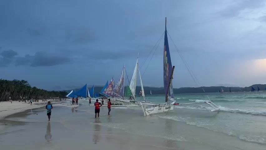 This video captures a beach scene at dusk, likely in the Philippines, given the presence of paraw sailboats. The sky is overcast and transitioning from daylight to twilight, with a hint of warm light on the right horizon. The water is a clear turquoise, with gentle waves lapping onto a white sandy beach. In the foreground, three individuals are visible walking on the wet sand near the water's edge. One person is on the far left, wearing a blue t-shirt and dark shorts, walking towards the viewer. In the middle ground, two people, one in a red t-shirt and black shorts and another in a red shirt and dark shorts, are standing and looking towards the water. To their right, another person is partially visible, bent down near the water. A line of paraw sailboats, each with a distinctive outrigger design and colorful sails, are anchored or beached along the shoreline, extending into the distance. The sails are mostly furled, but some display vibrant patterns. In the far left background, palm trees line the edge of the beach, and the silhouettes of more sailboats can be seen further down the coast. The overall atmosphere is serene, with the gentle sounds of the waves and the approaching evening.