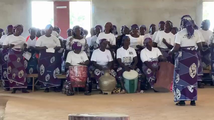This media file shows a group of women, likely a choir or a religious group, gathered indoors. The subjects are predominantly women dressed in matching white tops and vibrant purple and white patterned skirts. They are arranged in rows, with some standing and others seated. Several women in the front are actively engaged in playing percussion instruments: two are playing drums, and one is playing a large gourd-like instrument. The others appear to be singing or about to sing, with some holding papers.

The setting appears to be a large, open room with bare concrete walls and a light-colored floor. Two large windows are visible in the background, letting in natural light, suggesting it's daytime. The overall atmosphere seems energetic and celebratory, indicated by the women's engaged postures and facial expressions. The presence of musical instruments and the organized arrangement of the group point to a performance or a religious service.

Notable details include the distinct uniforms of the women, the traditional-looking drums, and the large, decorative gourd instrument. The scene takes place in Kufana, Nigeria. There is some visible text on the back of one woman's shirt, but it is too small and blurry to be legible. The lighting suggests it is daytime, and the indoor setting makes weather irrelevant. The emotions conveyed are positive, suggesting a sense of community and shared purpose.