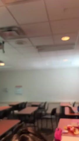 The video shows a blurred classroom scene with students sitting at desks. The camera moves and focuses on a student with her hair pulled back in a bun. The walls are white and light blue. There are other students in the room, some with laptops. The room is lit by overhead fluorescent lights.