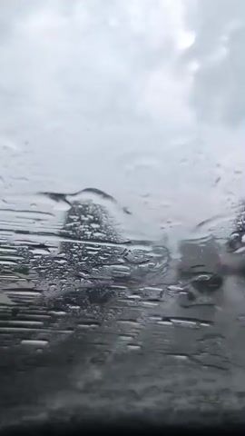 A POV shot from inside a car during heavy rain. Water droplets distort the view of a traffic scene. Cars are visible ahead, some with red taillights illuminated. The sky is overcast and gray. The windshield wipers are not actively clearing the rain, and the view is significantly obscured by the water on the glass.