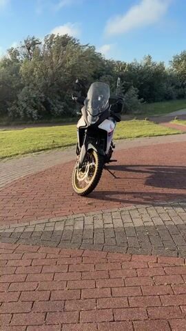 A white adventure motorcycle is parked on a brick path. To its right, a black cruiser motorcycle is parked. Further to the right, a blue street bike is parked. In the background, there is a grassy area with trees, a sidewalk, a bench, and some buildings. The sky is blue with some clouds.