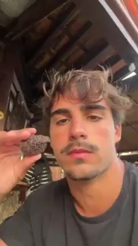 Here's a detailed description of the video:

A young man with fair skin, brown hair with light streaks, and a mustache is holding up a dark brown, circular object. He brings it to his mouth, takes a bite, and chews. The camera is close to his face, capturing his expressions as he eats.

He is sitting in what appears to be a cafe or outdoor restaurant setting. The background shows wooden beams, decorative items, and a cafe counter with a person wearing a striped shirt standing behind it.

The man seems to be enjoying what he is eating, focusing on the flavor.