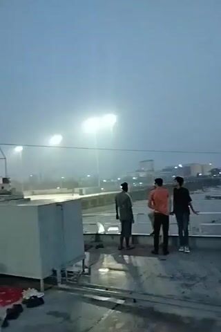 Here is a detailed description of the video:

The video appears to have been shot from a vessel, likely a ferry or large boat, as suggested by the view of the deck and the sound of a horn or engine. The setting is outdoors, on an overcast day with fog or mist obscuring the distance.

The focus is on the deck of the vessel where a number of people are present. Some are standing and looking out towards the shore or docks, while others are lying down on blankets and makeshift bedding, seemingly resting. 

The background shows a dock or port area with construction equipment, possibly a crane or large machinery, and buildings. There are also bright lights illuminating the area, cutting through the fog.

The overall impression is a scene of transit or waiting, with people in a transitional space. The fog and the presence of bedding suggest a long journey or a prolonged wait in less-than-ideal conditions.