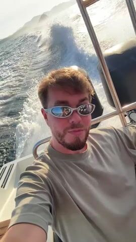 The video shows a selfie of a man with light brown hair and a beard, wearing a light green t-shirt and futuristic-looking silver sunglasses. He is on a boat, with the camera facing him. The boat is moving quickly, creating white waves in the blue water behind him. The sun is shining brightly, and there are islands visible in the distance. The side of the boat and a motor are also visible in the frame.