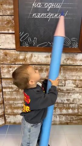 A young boy stands holding a large blue pencil in front of a chalkboard. The chalkboard has writing on it and drawings of school supplies. The boy is wearing a dark sweatshirt and jeans. He is looking at the chalkboard and holding the pencil with both hands. The background is made of wooden planks. There is a number "5" on the floor in front of the boy.
