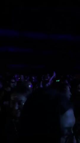 The video shows a crowd of people at a concert or music festival. The scene is lit with predominantly blue lights, creating a dynamic and energetic atmosphere. In the foreground, several individuals are visible from behind, their faces partially obscured by the angle or the lighting. One person in the immediate foreground has dark, curly hair. Further back, the crowd is dense, with many people raising their hands or holding up phones. The lighting is dramatic, with beams of blue light cutting through the darkness and illuminating the crowd and the stage area. On the stage, there are bright lights and what appears to be a large, sculpted figure or a stage design that resembles a head or a statue. The overall impression is that of a lively event with a focus on the performance and the collective experience of the audience.