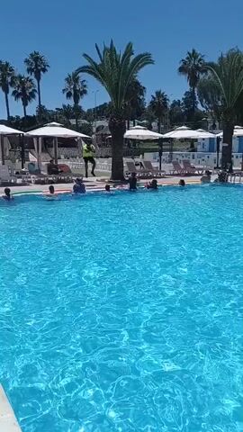 A group of people are in a swimming pool. A man in a yellow shirt and black shorts is standing on the edge of the pool and appears to be leading some sort of activity, possibly a water aerobics class. Several people are in the water, some on inflatable rings, and seem to be following his instructions. Palm trees and white umbrellas are visible in the background, suggesting a tropical or resort setting. The water is a bright blue and appears clear. The sky is clear and the sun is shining, indicating a bright, sunny day.