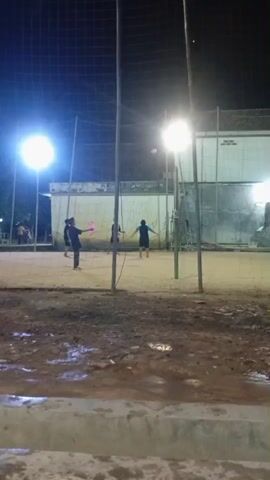 The video shows an outdoor volleyball court at night. Several people are playing volleyball under bright artificial lights. The court appears to be made of dirt or sand, and there are puddles of water on the ground, suggesting it may have rained recently. A net is set up for the game, and volleyball poles are visible. In the background, there is a building and some trees. The overall atmosphere is casual and recreational.