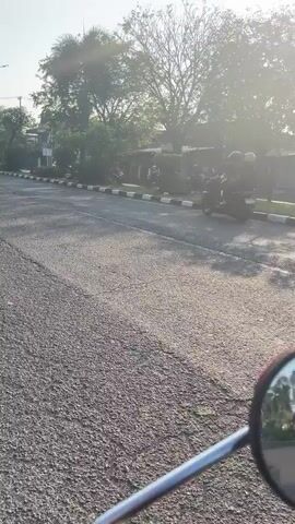 The video captures a street scene from the perspective of someone riding a motorcycle. In the foreground, the handlebars and a portion of the motorcycle's mirror are visible. The road surface is asphalt with visible cracks. A single motorcycle is moving away from the camera on the right side of the road, heading towards the distance. To the left of the road, there's a sidewalk with black and white striped curbing. Beyond the sidewalk are trees and buildings. The lighting suggests it is either early morning or late afternoon, with sunlight casting a soft glow and creating a hazy effect. The environment appears to be an urban or suburban area.
