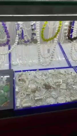 The camera pans across a jewelry display case. The case is filled with various silver and pearl jewelry items, including necklaces, pendants, and rings. The jewelry is arranged neatly in separate sections of the display case. The case has a glass top and interior lighting that illuminates the jewelry.