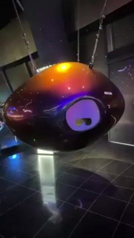 A still shot shows a large, eye-shaped structure suspended from the ceiling. It is metallic and reflective, with a color gradient from orange-gold at the top to purple-brown at the bottom. There is a white oval recess with visible 3D glasses inside. The structure hangs by thin cables above a black tiled floor. Reflections are visible in the structure's surface and on the floor. A section of a dark room with a wall display is visible in the background.