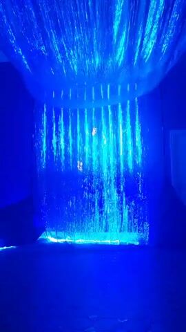 The video shows a dark room illuminated by blue lights. Hanging from the ceiling are strands of what appear to be fiber optic cables, emitting a bright blue light and creating a curtain-like effect. Below this, a sheet of translucent material also emits a blue light, with water cascading down its surface, creating a shimmering waterfall effect. The floor is dark and reflects the blue light.