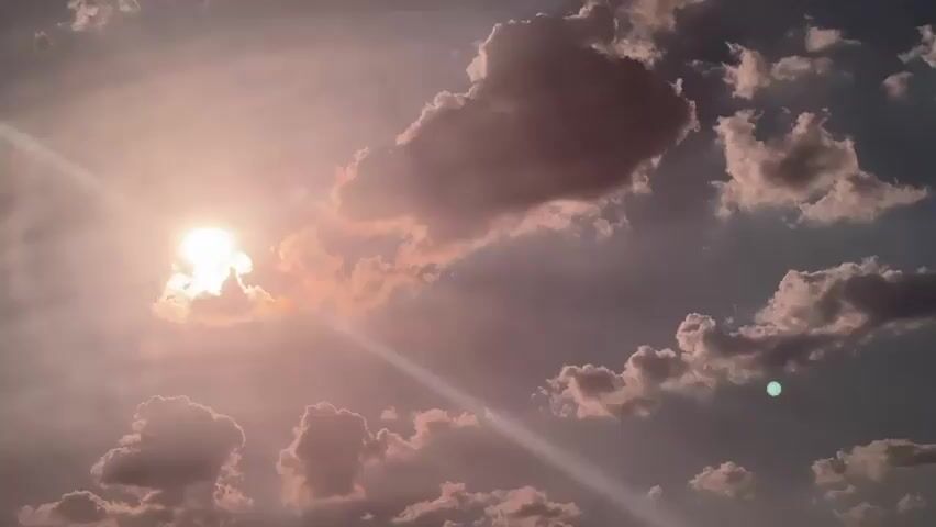 This media file features a sky filled with dramatic clouds during what appears to be sunset or sunrise, given the warm, golden light illuminating the scene. The sun, a bright orb, is partially obscured by clouds, casting strong rays that pierce through the atmosphere. The clouds themselves vary in texture and color, ranging from soft, fluffy white to deeper, more shadowed grays and pinks, suggesting a dynamic weather system.

A prominent object in the frame is a traffic signpost on the right side of the image. It has at least two signs: a circular one at the top and a rectangular one below it, which displays a green circular light. This suggests the location is near a road or intersection. A hint of a dark structure, possibly a building or roofline, is visible at the very bottom right corner.

The overall mood is serene and awe-inspiring, with the powerful presence of the sun and the textured clouds creating a visually striking panorama. The light suggests it is either early morning or late evening. The location context provided identifies Zheleznik, Bulgaria, as the setting. No discernible text is visible in the media itself.