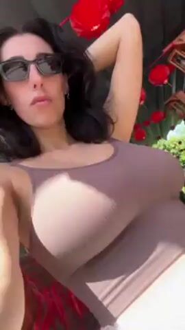 A woman with dark hair and sunglasses is taking a selfie. She is wearing a brown tank top and white shorts. The background includes red lanterns and greenery. The camera is held at a low angle, looking up at the woman. She adjusts her hair. The setting appears to be outdoors on a sunny day.