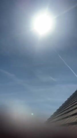 The video begins with a bright, sunny sky, revealing wispy clouds and a contrail from an aircraft. A roof with brown tiles is visible in the lower right corner. The camera then pans downward, showcasing a residential neighborhood with trees, houses, and hedges. The trees are predominantly green, and the houses have white walls and brown roofs. The sky remains clear and blue throughout the pan.