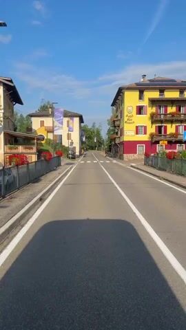 The video shows a view from a vehicle moving forward on a two-lane road lined with buildings and trees. The road has white lane markings and a pedestrian crossing. On the left, there is a building with a balcony adorned with red flowers, and banners with yellow and purple designs are visible. On the right, a yellow building with red accents and balconies is prominent, with solar panels on its roof. A person on a bicycle is seen cycling away from the camera in the middle of the road. The sky is clear and blue.