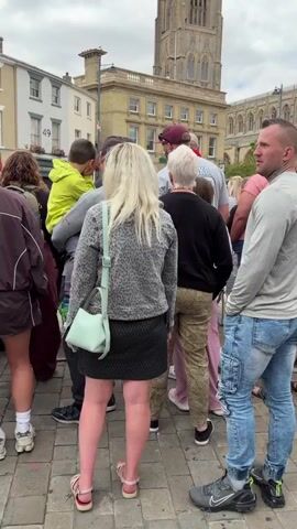 The video shows a crowd of people gathered outdoors. In the background, a large, ornate building with a tall spire, possibly a church or cathedral, is visible. Some people are wearing t-shirts with graphics, including one with the text "YOU AGAINST YOU" and a logo. A banner with images of muscular men and the text "MAYHEM GYM" is also present. The overall atmosphere appears to be casual, with people standing around and some engaging in conversation. The ground is paved with cobblestones.