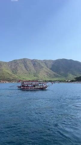 A medium-sized boat carrying passengers sails on the blue water of the sea. The boat is red and white with a white awning and multiple decks. The boat is moving from left to right in the frame. In the background, a lush green mountain range rises behind a beach lined with people and small buildings. The sky is clear blue with a few wispy clouds. The video shows the boat moving through the water with the mountainous coastline in the distance.