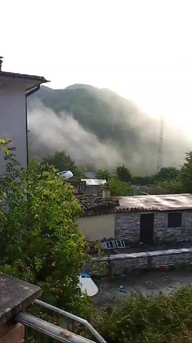 The video pans across a misty mountainous landscape with houses in the foreground. Sunlight streams through the mist, illuminating the peaks. There are trees and vegetation scattered throughout the scene, and a tall communication tower can be seen in the distance. The overall atmosphere is serene and tranquil.