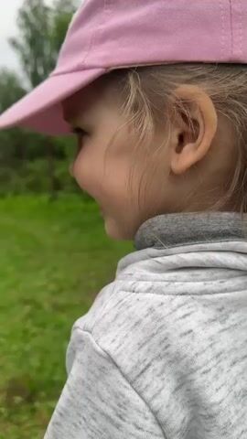 A young child wearing a pink cap, a grey hooded sweatshirt, and grey pants runs along a grassy path. The child is wearing black rubber boots and is moving away from the camera towards a dark-colored car parked in the distance. To the left of the path is a wooden fence, and to the right are trees and foliage. The sky appears overcast.