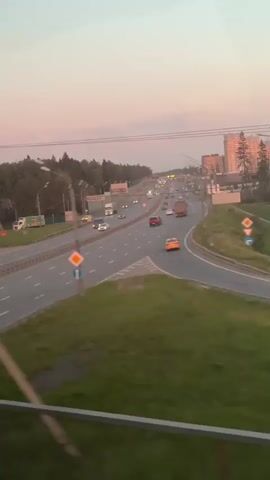The video shows a multi-lane highway with vehicles traveling in both directions.  A grassy median divides the lanes.  There are buildings in the distance that are catching the pink light of the sunset. There are electrical poles visible.  The video is shot from an elevated position.