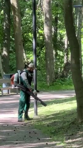 A man wearing a green uniform and a cap is using a leaf blower in a park. He has a backpack blower on his back and is holding the nozzle with both hands. He is facing to his right, blowing leaves on the ground. The man is standing on a dirt path next to a grassy area. There are trees in the background. A bench and a trash can are visible to the left of the man. The video is taken from a slightly low angle.