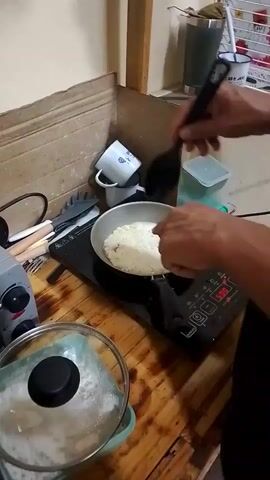 A person is cooking something in a frying pan on a black portable induction cooktop. The person holds a black slotted spoon in their hand. The countertop is made of light-colored wood planks. A glass pot lid sits on the counter. In the background, there is a wall with tiled stone texture. There is a white mug and other cooking utensils on the counter. A window can be seen in the background with a white shelf.