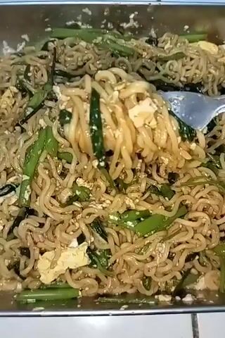 The video shows a close-up, overhead view of a serving of instant noodles mixed with vegetables and scrambled egg. A fork is lifting a portion of the noodles, revealing their texture and the other ingredients. The noodles are a light brown color, coiled and slightly greasy. Mixed in are green vegetables, appearing to be a leafy green like water spinach, cut into short segments, and pieces of scrambled egg that are pale yellow. The dish is served in a metal rectangular tray. The background is out of focus but appears to be a tiled surface. The lighting is bright, highlighting the food. The action is solely focused on the food and the fork interacting with it.