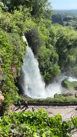 The video shows a broad, steep waterfall flowing downwards from left to right, surrounded by lush green trees and vegetation. At the base of the waterfall, a walkway with a low stone wall is visible, where several people are walking. The waterfall creates a misty spray as it crashes into the pool below. Beyond the immediate area of the waterfall, a wide, open landscape extends to the horizon under a clear, blue sky. The landscape appears to be a mix of cultivated fields and wooded areas. The camera pans slightly to the right, revealing more of the valley beyond the waterfall.