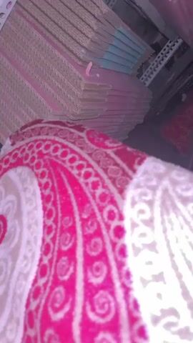 The media file shows a close-up view of a patterned textile, possibly a blanket or rug, in the foreground. The textile features a prominent paisley design in vibrant pink and cream colors. In the background, several stacks of flat, rectangular objects are visible, appearing to be cardboard boxes or similar packaging materials. These stacks are organized on white metal shelving units. The lighting suggests an indoor setting, possibly a storage area or a retail space. The overall impression is one of organized storage or perhaps a retail display of items. There is no visible text in the media file.