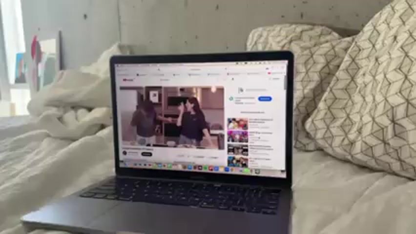 A laptop is resting on a white bed, displaying a YouTube video. The video appears to be a cooking segment featuring two people in a kitchen. The bed has white bedding and patterned pillows. A picture frame can be seen in the background to the left.