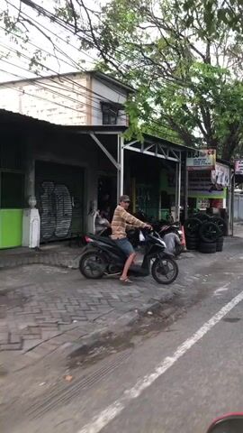 A man on a black scooter, wearing a striped shirt and blue shorts, is parked on a sidewalk in front of a small shop. The shop appears to be a tire repair business, with stacks of tires visible outside. Inside the shop, several people are working on a motorcycle. One person is kneeling and appears to be fixing a tire. Another person is standing and observing. The environment is an urban street with buildings and trees. The lighting suggests it is daytime.