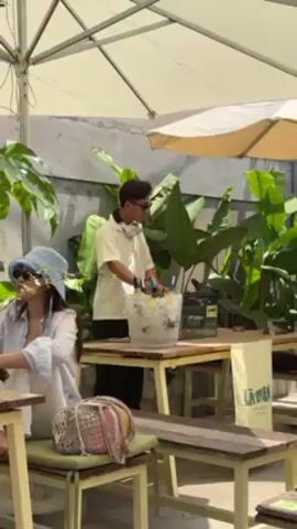 Here is a concise description of the video:

The scene is an outdoor cafe or patio setting. A woman with a blue bucket hat and sunglasses sits at a wooden table with a cushion, seemingly occupied with something on the table in front of her. In the background, a man wearing sunglasses and headphones is arranging bottles in an ice bucket placed on another wooden table.  Green plants provide a backdrop, and shade is provided by umbrellas overhead. There is also a sign on the back of one of the chairs reading "LÀMEN".