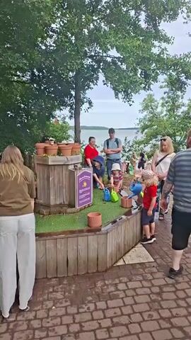 A group of people, including several children and adults, are gathered around a wooden planter box with several terracotta pots on top. The planter box has a sign on it with purple text and an illustration of a watering can. Some of the children are holding small watering cans and appear to be watering the plants or the grass-like covering on the planter. The scene is outdoors, with trees and a body of water in the background. The ground is paved with brick. The overall atmosphere appears to be one of a family-friendly outdoor activity or event.
