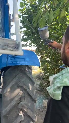 A person is spray painting a blue tractor. The person is holding a spray gun and is wearing a black shirt and a green and white scarf around their neck. The tractor is large and has a blue body and a large black tire. The tractor is parked outdoors in a grassy area with trees in the background. The sky is clear and blue. There is some paint overspray visible on the ground around the tractor. The person is painting a white part of the tractor.
